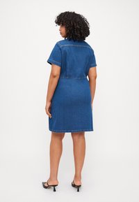 Denimkjole i mørkeblå, kortermet med krage, med sømadetaljer. Den faller over kneet, og er parret med svarte hæl sandaler.