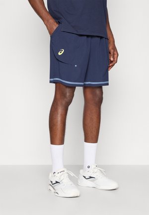 Pantalones cortos deportivos en color navy con textura de malla, bolsillos laterales y una franja de acento en azul claro. Se llevan con calcetines blancos y zapatillas blancas.