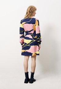 Buntes Kleid mit navy-blauem Hintergrund, verziert mit abstrakten Mustern in Pink, Gelb und Grün. Das Kleid hat lange Ärmel und eine taillierte Passform. Dazu schwarze Knöchelstiefel.