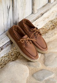 Brown leather shoes with round toes, rubber soles, woven jute detailing, and laces. The tongue features a small embossed logo.