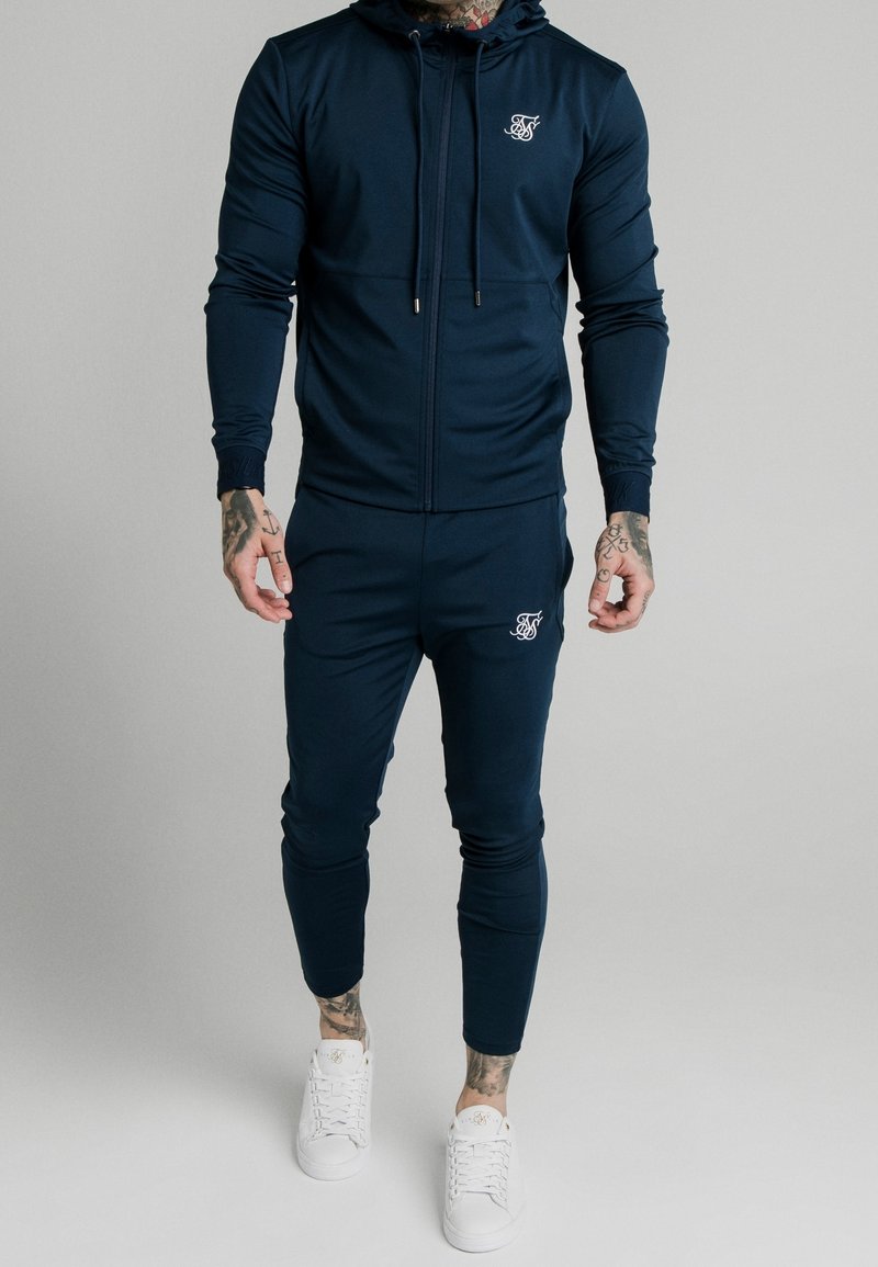Homme portant un sweat à capuche zippé bleu marine et un jogging assorti avec des logos blancs, des baskets blanches et des tatouages visibles sur les mains et les chevilles.