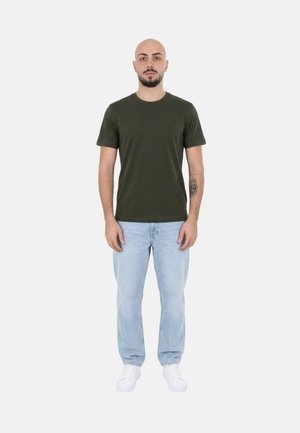 Uomo con barba e tatuaggio in piedi, indossa una maglietta verde scuro, jeans azzurri e scarpe da ginnastica bianche su uno sfondo uniforme.