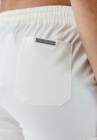 Weiße Shorts mit einem elastischen Bund, die ein rechteckiges Metalletikett an der Gesäßtasche haben. Glattes Material mit matter Oberfläche.