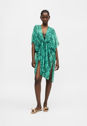 Žena má na sebe zelený vzorovaný priehľadný prehoz previazaný v páse cez čierne plavky, stojí bosá s béžovými sandálmi na bielom pozadí.