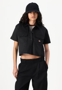 Camisola preta cropped com mangas curtas, dois bolsos no peito e um pequeno logotipo. Combinada com calças pretas de cintura alta e um boné correspondente.