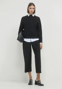 Maglione nero a maglia sopra una camicia bianca con colletto, abbinato a pantaloni neri cropped. Modella che indossa scarpe con tacco quadrato nere lucide, mentre tiene una borsa nera.