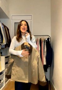 Woman in white long-sleeve holds tan jacket with dark collar on hanger in a small room with clothes rack and shelves.