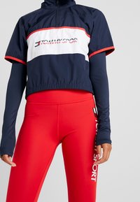 Marinefarbener, taillierter Blazer mit weißen und roten Akzenten, der das "TOMMY SPORT"-Logo trägt. Kombiniert mit enganliegenden roten Leggings, die das Logo vertikal anzeigen.