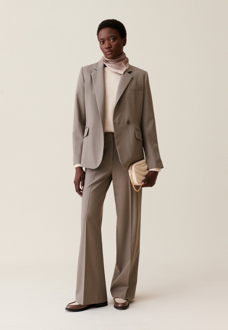 Szary, dopasowany garnitur z usztywnioną marynarką i rozszerzającymi się spodniami, zestawiony z kremowym swetrem i jasnoróżowym szalikiem, trzymając kremową torebkę.