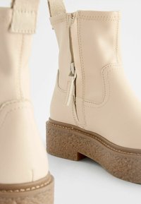 Beige Knöchelstiefel aus glattem Synthetikleder mit einer strukturierten braunen Plateausohle und einem seitlichen Reißverschlussdetail.