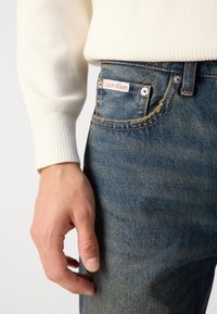 Jeans de mezclilla azul claro con un sutil desgaste y costuras naranjas en contraste, que presentan un logo de Calvin Klein en rojo y blanco en el bolsillo frontal.