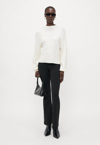 Pull blanc, pantalon noir ajusté, bottines à talons noires. Le modèle tient un petit sac noir. Les accessoires comprennent des lunettes de soleil noires et des boucles d'oreilles créoles.