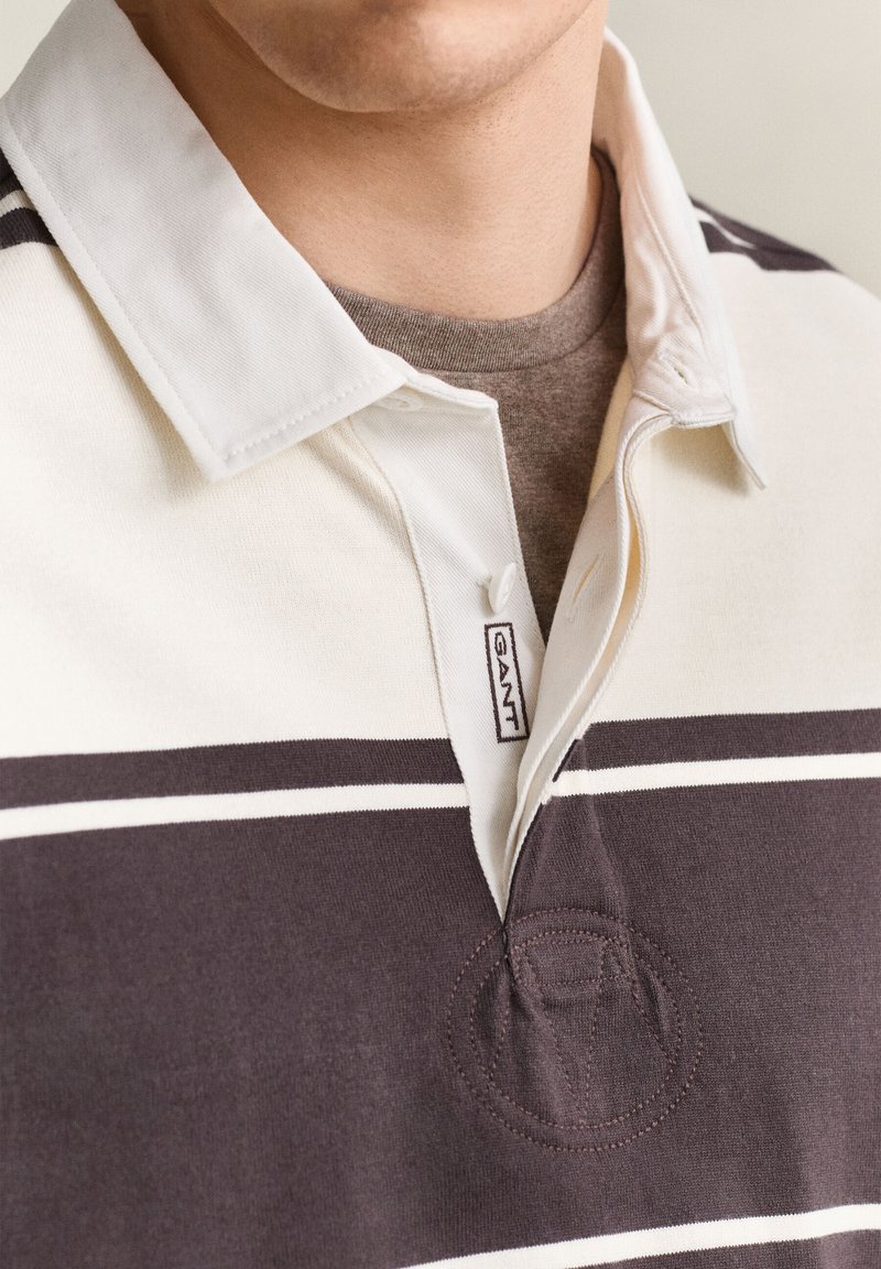 Close-up of a person wearing a brown and white striped polo shirt with a white collar and a visible GANT brand tag.