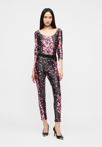 Γυναίκα που φοράει ροζ και μαύρη ολόσωμη φόρμα με μοτίβο, με μανίκια τρία τέταρτα και μαύρα ψηλά τακούνια, στέκεται μπροστά σε απλό φόντο.