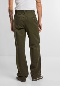 Olijfgroene katoenen broek heeft een ontspannen pasvorm, twee achterzakken met knopen en een soepele textuur. Gecombineerd met sportieve schoenen.