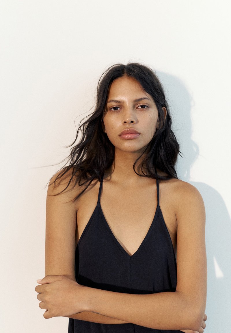 Femme aux longs cheveux foncés portant un haut noir sans manches, debout, les bras croisés, devant un fond clair uni.