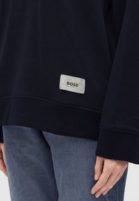 Personne portant un sweat-shirt noir avec une étiquette "BOSS" près de l'ourlet et un pantalon en jean bleu, main reposant le long du corps.