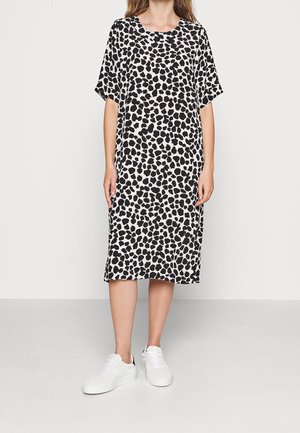 Kvinde iført en løs sort og hvid plet midi-kjole med korte ærmer og hvide sneakers, stående mod en ensfarvet baggrund.