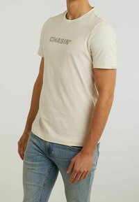 Vaaleanbeige lyhythihainen T-paita, jossa on painettuna "CHASIN'" hieman tummemmalla sävyllä. Sisältää pyöreän kaula-aukon ja kevyen, sileän kankaan rakenteen.