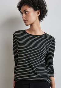 Junge Frau mit kurzen lockigen Haaren, die ein schwarz-weiß gestreiftes Langarmshirt trägt und nach links gegen eine schlichte Wand schaut.