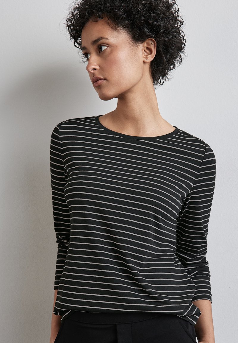 Junge Frau mit kurzen lockigen Haaren, die ein schwarz-weiß gestreiftes Langarmshirt trägt und nach links gegen eine schlichte Wand schaut.