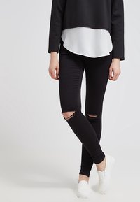 Jeans negros ajustados con desgastes en las rodillas. El modelo lleva un sudadera negra corta sobre una camiseta blanca. Zapatillas blancas sin cordones completan el look.