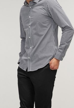 Mężczyzna w czarno-białej koszuli w kratę zapinanej na guziki i czarnych spodniach, z jedną ręką w kieszeni, na jednolitym tle.