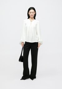 Blouse blanche en satin à manches bouffantes et col en V, associée à un pantalon noir taille haute. Sac à main noir et chaussures à bouts pointus.