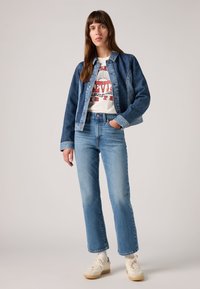 Veste en jean bleu foncé, t-shirt graphique blanc, jeans droite bleu clair, baskets crème, modèle se tient contre un fond neutre.