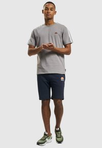 Camiseta gris de manga corta con franjas contrastantes en los hombros, combinada con pantalones cortos azul marino que presentan un logo. Llevado con zapatillas deportivas verdes y blancas.