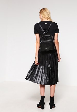 Femme portant un haut noir à manches courtes, une jupe plissée en cuir noir, des bottines noires et un sac à dos texturé noir, se tenant de dos.