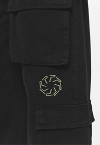 Svart tyg cargo byxor med en stor sidoficka och en cirkulär grön broderad logotyp med ett geometriskt mönster. Texturen är slät.