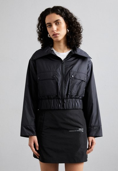 Veste noire cintrée avec grand col et poches avant, dotée d'une fermeture éclair et d'une taille élastique, associée à une mini-jupe noire.