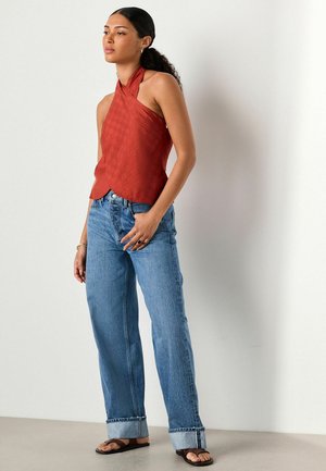 Frau mit lockigem Haar in einem rostfarbenen Neckholder-Top, hochgekrempelten blauen Jeans und braunen Sandalen, die mit einer Hand in der Tasche vor einer weißen Wand steht.