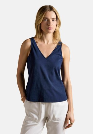 Žena se světlými vlasy, oblečená v tmavě modrém tílku s výstřihem do V a bílých kalhotách, stojící s jednou rukou v kapse na bílém pozadí.