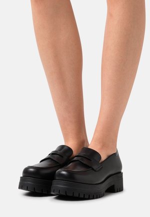 Mocassins en cuir noir avec un bout rond, dotés de deux larges brides et d'une semelle en caoutchouc épaisse avec des détails de texture.