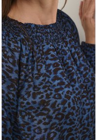 Blue animal print top with a smocked neckline and long sleeves, featuring a textured fabric and contrasting black leopard pattern.