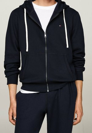 Mężczyzna ubrany w granatową bluzę z kapturem na zamek błyskawiczny z białymi sznureczkami oraz dopasowane granatowe spodnie, pod bluzą widoczna biała koszulka.