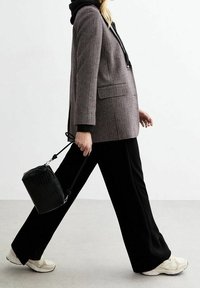 Blazer à carreaux avec une capuche noire, coupe oversize, pantalon large noir et un petit sac noir texturé. Des baskets beiges complètent la tenue.