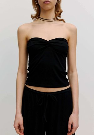 Vrouw draagt een zwart strapless top en zwarte broek met trekkoord, gecombineerd met creolen en een gelaagde zwarte chokerketting.