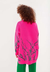 Överdimensionerad rosa tröja med ett grönt abstrakt mönster, långa ärmar och ribbat avslut. Bärs med gröna byxor, texturen verkar mjuk.