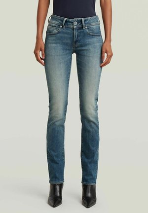 Femme portant un jean skinny bleu délavé et des bottines noires à bout pointu, debout devant un fond uni.