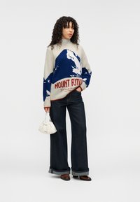 Πλεκτό πουλόβερ σε εκρού και μπλε με γραφικό βουνού και το κείμενο «MOUNT FUTURE», συνδυασμένο με παντελόνι από σκούρο τζιν με φαρδιά μπατζάκια και λευκή τσάντα.