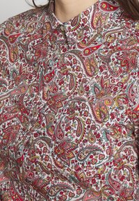 Gros plan sur un col de chemise boutonné avec un motif floral cachemire détaillé rouge, jaune et vert sur fond blanc.