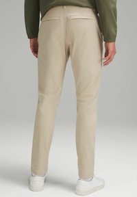 Beżowe spodnie z lekkiego materiału, slim fit, z tylnymi kieszeniami. W połączeniu z białymi sneakersami i zieloną bluzką. Gładka tekstura.