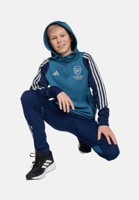 Blauwe hoodie met rits, Arsenal-logo en de tekst "Emirates Fly Better". In combinatie met navy trainingsbroek en zwarte sneakers met witte accenten.