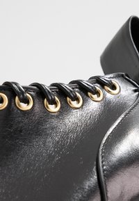 Zapato de cuero negro con ojales dorados, cordones negros y una textura suave con detalles de costura mínimos en la parte superior.