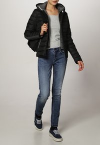 Sort pufferjakke med grå indvendig, grå langærmet top, blå denim jeans og navy sneakers, gående mod en lys baggrund.