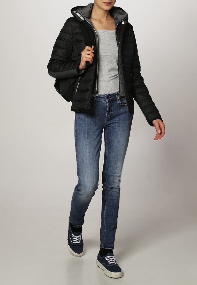 Sort pufferjakke med grå indvendig, grå langærmet top, blå denim jeans og navy sneakers, gående mod en lys baggrund.