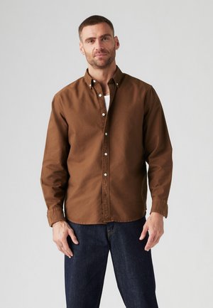 Camisa marrón de botones, fabricada en algodón, de corte relajado, con mangas largas y puños abotonados, combinada con jeans de mezclilla azul oscuro. Diseño sencillo, sin patrones.